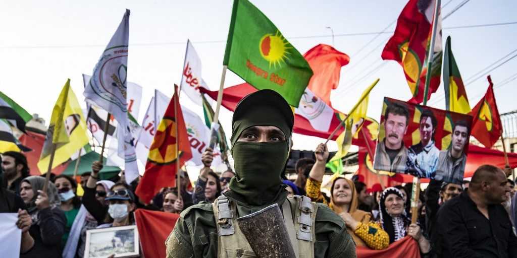 In the front row, a masked soldier stands holding his weapon against his chest. In the back, a row of Kurdish flags, banners and placards held by Kurdish protesters.