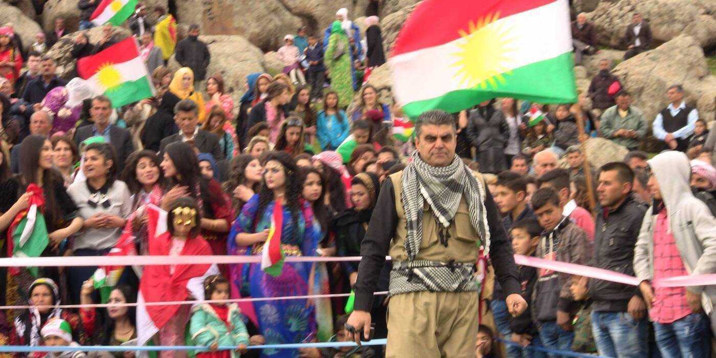 Barzan Hussein stands in front of a crowd, Kurdistan flags are visible in the background.