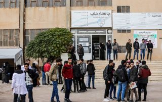 Students stand in front of the door of the university. Above the door, a sign in Arabic: "University of Aleppo"