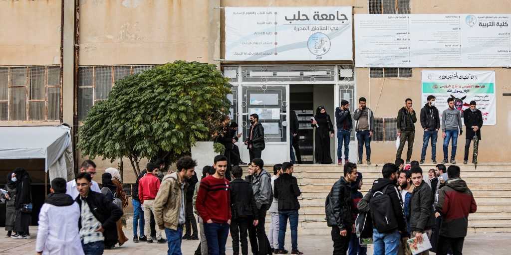 Students stand in front of the door of the university. Above the door, a sign in Arabic: "University of Aleppo"