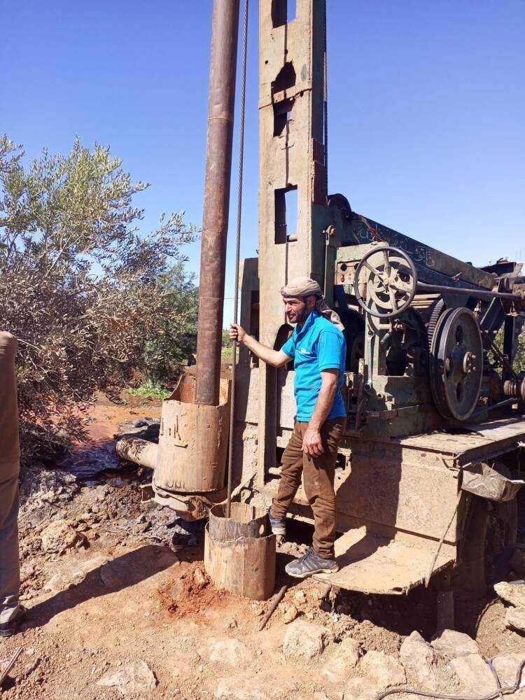 رجل يقف أمام حفارة آبار في مدينة إنخل شمال درعا، أثناء حفر بئر ضمن مبادرة شعبية لتوفير مياه الشرب، 06/ 04 / 2023 (مدينتي إنخل)