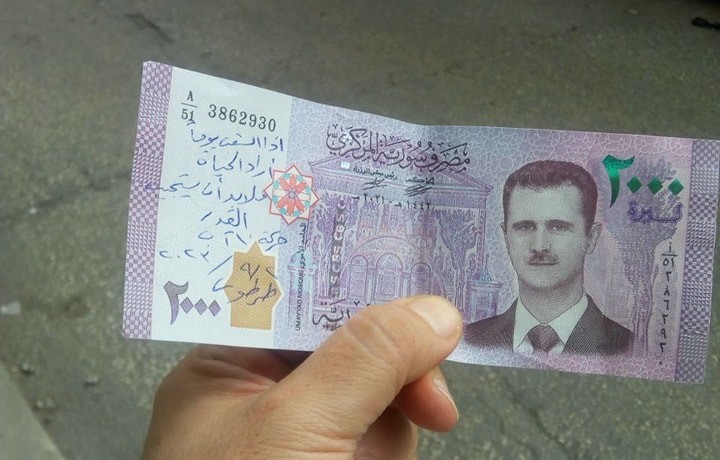 A member of the 10th of August Movement—an opposition movement launched in Syria’s regime-held coastal provinces in summer 2023—holds up a 2,000 Syrian pound bank note in Tartous city. The writing on the bill reads: “If the people one day wanted life, fate must respond,” 2/9/2023 (10th of August Movement)