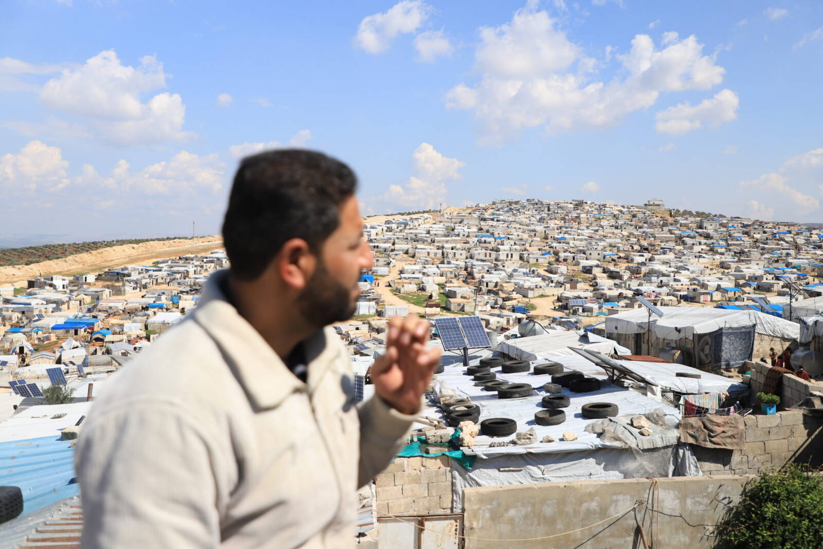 مازن الوراد، يقف على سطح منزله في مخيم "أم الشهداء" بأطمة على الحدود السورية-التركية، وتظهر في الصورة عشرات الوحدات السكنية في المخيم، 10/ 03/ 2024 (عبد المجيد القرح/ سوريا على طول)