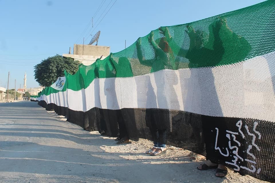 نساء يحملن علم الثورة السورية، طوله 75 متراً، خلال ورشة عمل للأشغال اليدوية في مركز مزايا النسائي بمدينة كفرنبل، عام 2014، (منظمة مزايا)