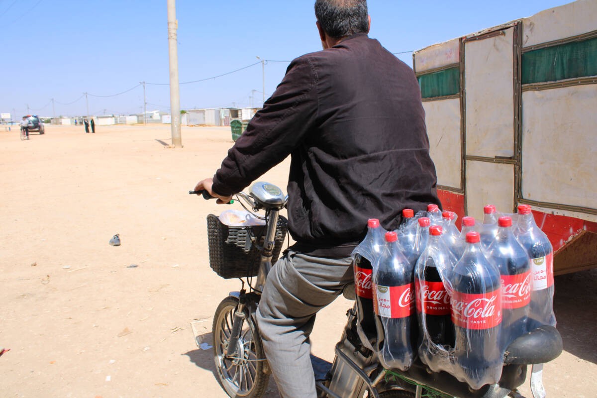 A Syrian man rides his bike loaded with two backs of Coca-Cola he purchased with WFP food assistance in Zaatari camp and intends to sell for cash, 3/4/2024 (Hanna Davis/ Syria Direct)