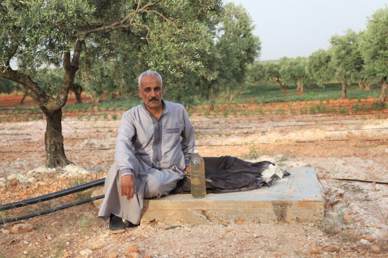 Khaled al-Jaabi sits at the edge of his well in Idlib’s al-Sanama village, showing a bottle of discolored water contaminated by the al-Hebat dump, 27/5/2024 (Abd Almajed Alkarh/Syria Direct)